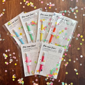 Op de afbeelding: Een verzameling feestartikelen, elk met een woordzoekpuzzel en een gekleurd potlood. De potloden zijn blauw, groen, geel, oranje en roze. De gunsten zijn verpakt in doorzichtige plastic zakken.