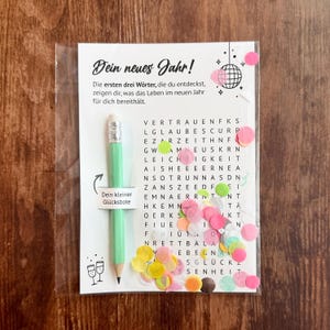 Op de afbeelding: Een doorzichtige plastic verpakking met een wenskaart met Duitse tekst en een lichtgroen potlood. De kaart heeft een woordzoekpuzzel met kleurrijke confetti en een discobal-illustratie. Het potlood heeft de tekst "Dein kleiner Gl&uuml;cksbote".