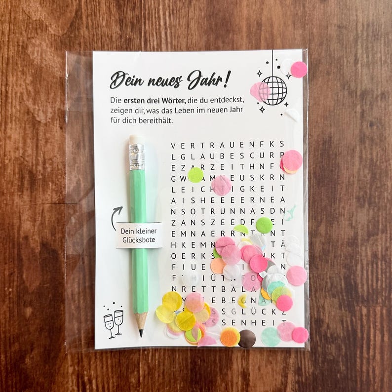 Op de afbeelding: Een doorzichtige plastic verpakking met een wenskaart met Duitse tekst en een lichtgroen potlood. De kaart heeft een woordzoekpuzzel met kleurrijke confetti en een discobal-illustratie. Het potlood heeft de tekst "Dein kleiner Gl&uuml;cksbote".