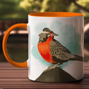May include: A white ceramic mug with an orange rim features a colorful illustration of a bird perched on a rock. The bird has a red breast, gray and brown wings, and a black beak.