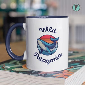 May include: White ceramic mug with a blue interior and handle. The mug features a blue whale illustration inside a circle with the words "Wild Patagonia" in a script font. The background of the circle has a sunset design.