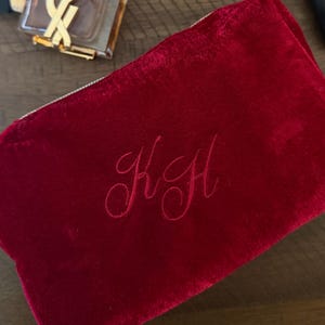May include: A deep red velvet zippered pouch featuring the embroidered monogram letters KH in a cursive script on the front surface. The pouch has a soft, plush texture and a rectangular shape with rounded corners.