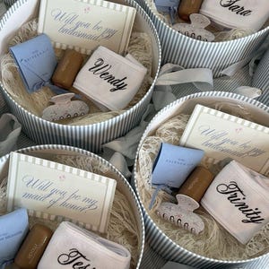 May include: Round gift boxes with gray and white striped sides containing a white headband with a name embroidered in black, a light blue pouch, a floral patterned hair claw, and a card reading Will you be my bridesmaid or maid of honor.