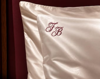 May include: A pair of cream-colored satin pillowcases with a smooth, reflective finish. The front pillowcase features an embroidered monogram in a dark burgundy script font with the letters FB, positioned in the upper left corner.