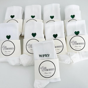 May include: White ribbed Pilates socks with a green heart embroidered near the top. Each pair is packaged with a cream-colored band featuring the text "EVER EMBROIDERED Pilates Socks" and sizes 5-10. One pair reads "WIFEY".