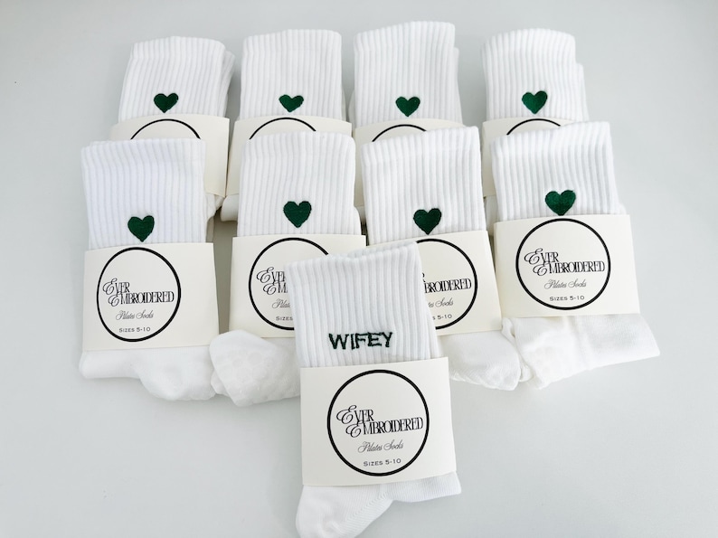 May include: White ribbed Pilates socks with a green heart embroidered near the top. Each pair is packaged with a cream-colored band featuring the text "EVER EMBROIDERED Pilates Socks" and sizes 5-10. One pair reads "WIFEY".