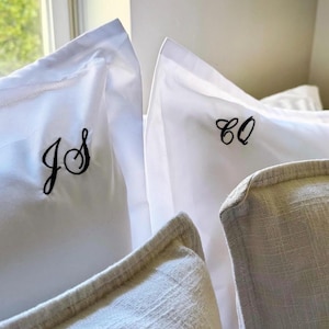 May include: Close-up of white pillowcases embroidered with black initials. One pillowcase displays "JS" and another "CO". The image also shows a textured, light-colored pillow.