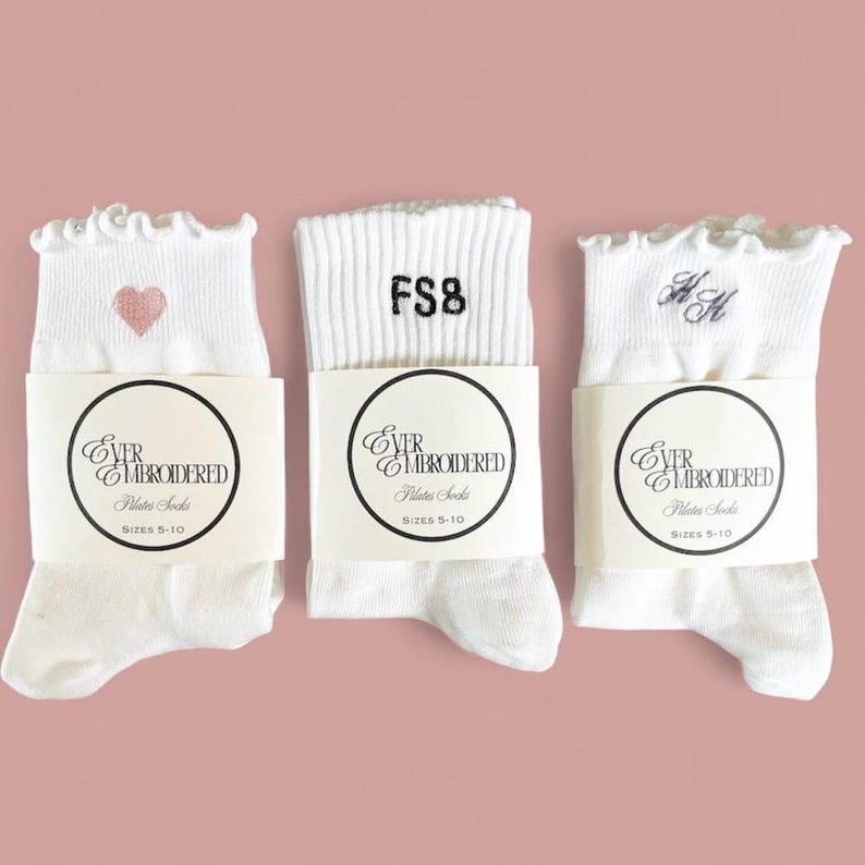 May include: Three pairs of white Pilates socks with ruffled tops, each wrapped with a label. One sock has a pink heart, another has "FS8" embroidered, and the last has embroidered initials. The labels read "EVER EMBROIDERED Pilates Socks Sizes 5-10."
