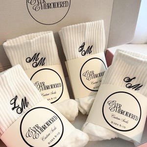 May include: Three pairs of white ribbed crew socks featuring black embroidered initials. Each pair is wrapped in a paper band labeled Ever Embroidered Custom Socks Sizes 5-10. The socks are presented inside a branded cardboard box.