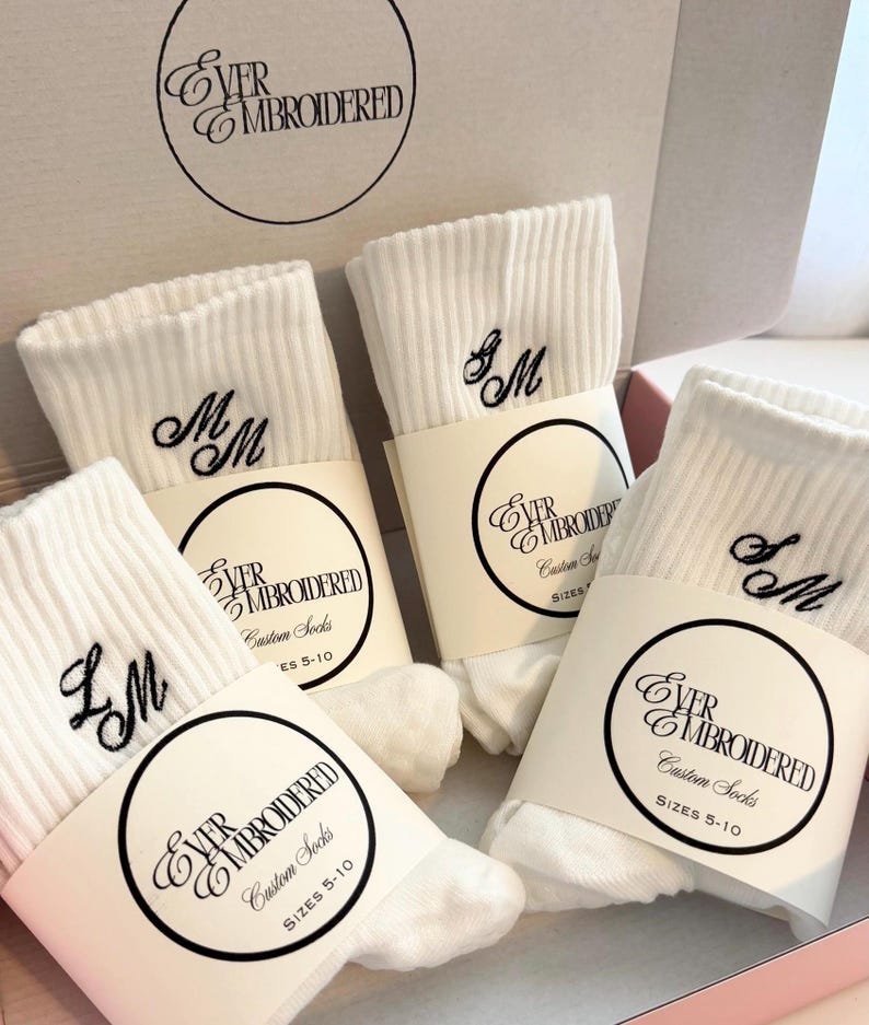 May include: Three pairs of white ribbed crew socks featuring black embroidered initials. Each pair is wrapped in a paper band labeled Ever Embroidered Custom Socks Sizes 5-10. The socks are presented inside a branded cardboard box.