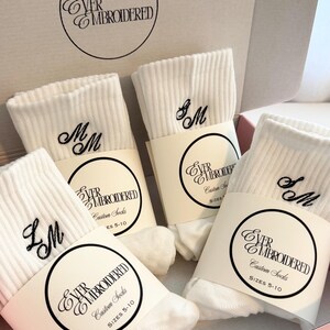 May include: White ribbed socks with embroidered initials, presented in a pink box. Each pair is wrapped with a label that reads "EVER EMBROIDERED Custom Socks" and sizes 5-10. The socks feature black embroidered initials.