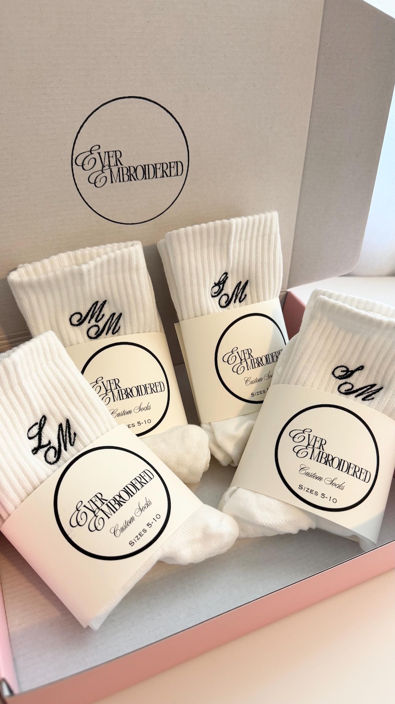 May include: White ribbed socks with embroidered initials, presented in a pink box. Each pair is wrapped with a label that reads "EVER EMBROIDERED Custom Socks" and sizes 5-10. The socks feature black embroidered initials.