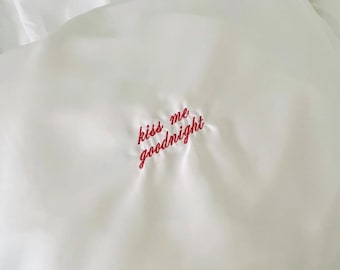 Pode incluir: Fronha de cetim branco com a frase bordada "kiss me goodnight" em escrita vermelha. A fronha está ligeiramente enrugada, sugerindo uma textura macia e confortável. A estética geral é simples e romântica.