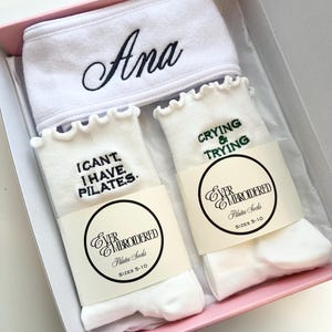 May include: A gift set in a pink box containing a white headband embroidered with the name "Ana" in black script, and two pairs of white Pilates socks. One pair of socks has the text "I CAN'T I HAVE PILATES" and the other pair has the text "CRYING & TRYING".