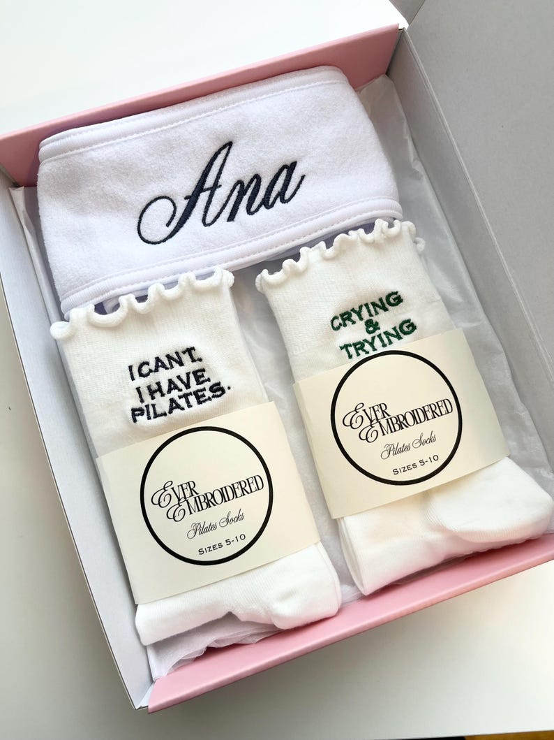 May include: A gift set in a pink box containing a white headband embroidered with the name "Ana" in black script, and two pairs of white Pilates socks. One pair of socks has the text "I CAN'T I HAVE PILATES" and the other pair has the text "CRYING & TRYING".