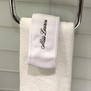 May include: A white, textured towel hanging on a chrome towel ring. The towel has the embroidered text "Miss Lauren" in black script. The towel is folded over the ring, ready for use in a bathroom setting.