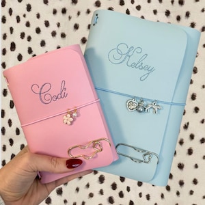 May include: A pink notebook personalized with the name Codi and a light blue notebook personalized with the name Kelsey. Both feature elastic bands with hanging charms, including a flower, pearl, heart, and leopard, plus large metal paper clips.