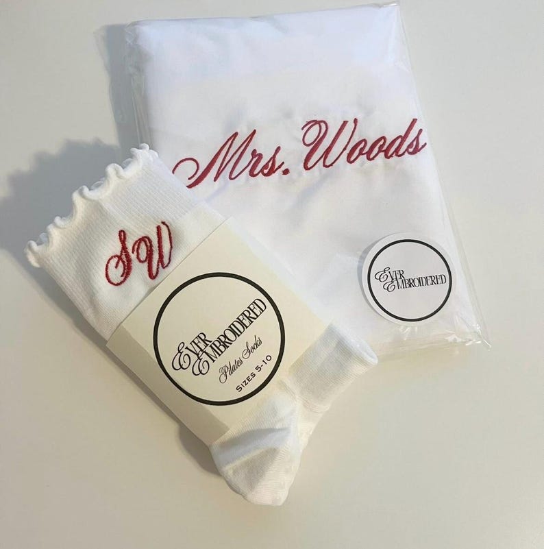 May include: White Pilates socks with a ruffled top and red embroidered initials "SW". A white pillowcase with "Mrs. Woods" embroidered in red is in a clear plastic bag. A round sticker with "Ever Embroidered" is also visible.