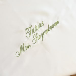 May include: Ivory-colored fabric with green embroidered text that reads "Future Mrs. Rozenboom" in a cursive font. The fabric has a subtle sheen, suggesting a satin or silk material. The embroidery is delicate and elegant.