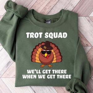 May include: Green sweatshirt featuring the text "TROT SQUAD" and a cartoon turkey wearing sunglasses and a hat. The text "WE'LL GET THERE WHEN WE GET THERE" is below the turkey graphic. The sweatshirt is folded on a wooden surface.