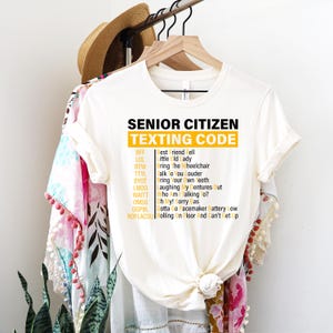 May include: White t-shirt with the text "SENIOR CITIZEN TEXTING CODE" in black and yellow. The shirt lists common texting abbreviations and their meanings, such as "LOL" for "little old lady."