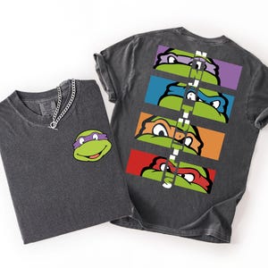 May include: A charcoal grey t-shirt featuring a small purple-masked turtle graphic on the front left chest. The back displays four horizontal rectangular panels, each showing a different coloured turtle mask, with the text NINJA TURTLES vertically.