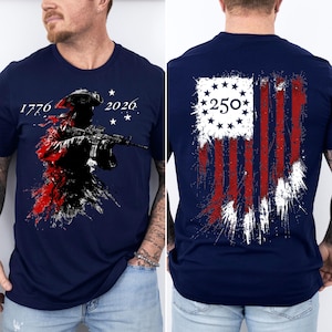 May include: A navy blue t-shirt featuring a front graphic of a soldier in a tricorn hat holding a rifle with the text 1776 and 2026. The back graphic displays a distressed American flag design with a circle of stars and the number 250 in the center.