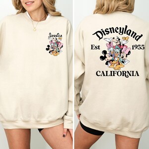 May include: Cream-colored sweatshirt featuring a Disney character graphic on the front and back. The front displays a cluster of characters with the name "Jessica" above. The back features "Disneyland Est 1955 California" with a character graphic.