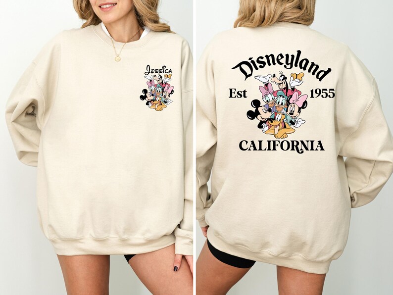 May include: Cream-colored sweatshirt featuring a Disney character graphic on the front and back. The front displays a cluster of characters with the name "Jessica" above. The back features "Disneyland Est 1955 California" with a character graphic.