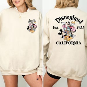 May include: Cream-colored sweatshirt featuring a Disney character graphic on the front and back. The front displays a cluster of characters with the name "Jessica" above. The back features "Disneyland Est 1955 California" with a group of Disney characters.