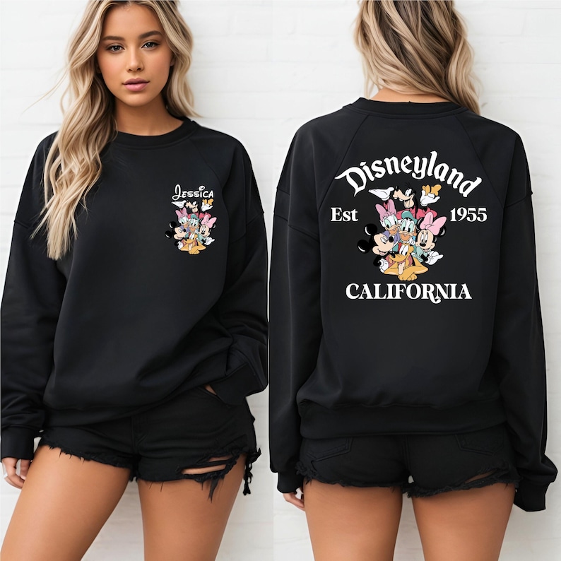 May include: Black sweatshirt featuring a colorful graphic of Disney characters. The front has the name "Jessica" above the graphic. The back displays "Disneyland Est 1955 CALIFORNIA" in white. The sweatshirt is worn with black shorts.