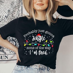 May include: Black t-shirt with the text "Probably just gonna jingle some of the way. I'm tired." The design includes a Santa hat, Christmas lights, and snowflakes. The shirt is a crew neck.