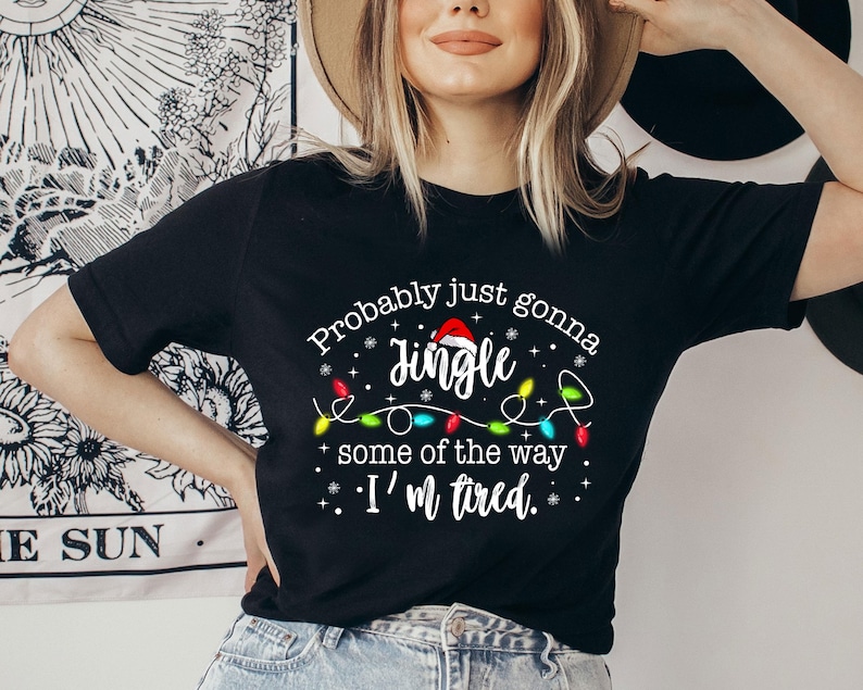 May include: Black t-shirt with the text "Probably just gonna jingle some of the way. I'm tired." The design includes a Santa hat, Christmas lights, and snowflakes. The shirt is a crew neck.