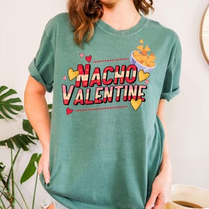 May include: A teal-colored t-shirt with the words "Nacho Valentine" in a playful font. The design includes red and yellow hearts, and a bowl of nachos. The shirt is a casual, comfortable style.