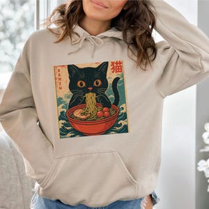 May include: Beige hoodie featuring a graphic of a black cat eating ramen from a red bowl. The cat has orange eyes and is surrounded by Japanese text and wave patterns. The hoodie has a front pocket and a drawstring hood.