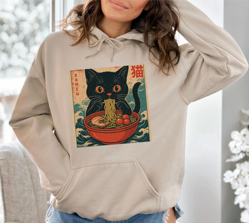 May include: Beige hoodie featuring a graphic of a black cat eating ramen from a red bowl. The cat has orange eyes and is surrounded by Japanese text and wave patterns. The hoodie has a front pocket and a drawstring hood.
