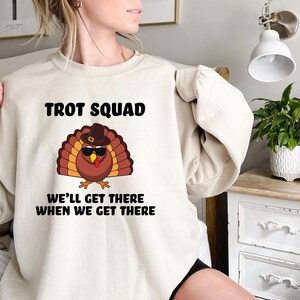 May include: A cream-coloured sweatshirt featuring the text "TROT SQUAD" above a cartoon turkey wearing sunglasses and a hat. Below the turkey is the text "WE'LL GET THERE WHEN WE GET THERE".