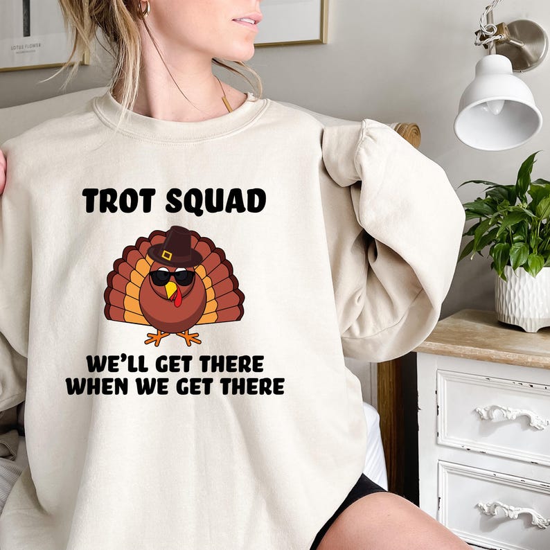 May include: A cream-coloured sweatshirt featuring the text "TROT SQUAD" above a cartoon turkey wearing sunglasses and a hat. Below the turkey is the text "WE'LL GET THERE WHEN WE GET THERE".