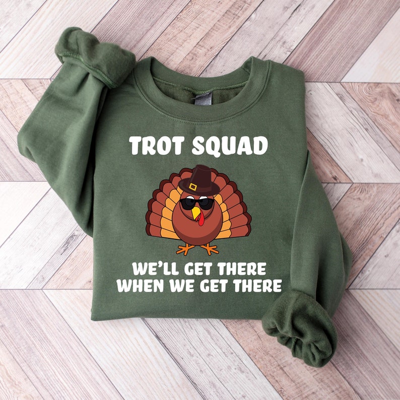 May include: Green sweatshirt featuring the text "TROT SQUAD" and a cartoon turkey wearing sunglasses and a hat. The text "WE'LL GET THERE WHEN WE GET THERE" is below the turkey graphic. The sweatshirt is folded on a wooden surface.