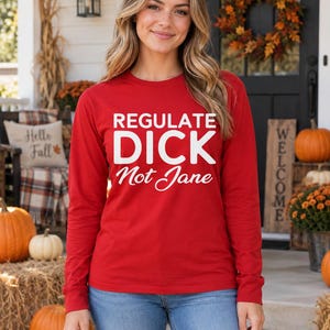 May include: A long-sleeve red shirt featuring white text that reads REGULATE DICK Not Jane in a combination of bold sans-serif and cursive script fonts. The garment has a crew neckline and straight hem, worn with blue denim jeans.
