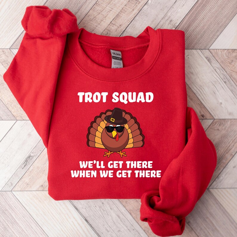 May include: A red sweatshirt featuring the text "TROT SQUAD" in white. A cartoon turkey wearing sunglasses and a hat is below the text. The phrase "WE'LL GET THERE WHEN WE GET THERE" is printed below the turkey illustration.