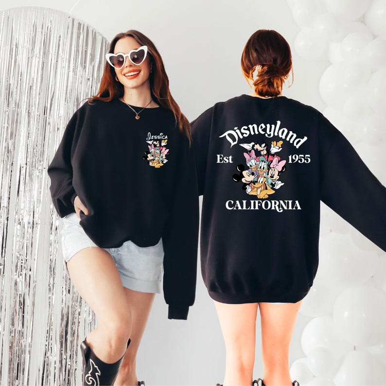 May include: Black sweatshirt featuring a Disneyland graphic on the back with the text "Disneyland Est 1955 California". The front of the sweatshirt has a smaller graphic with the name "Jessica". The sweatshirt has long sleeves.