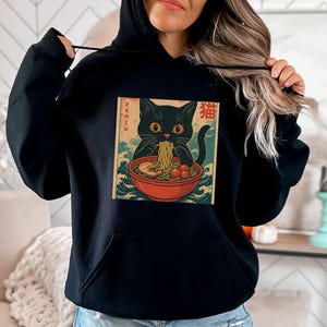 May include: Black hoodie featuring a graphic of a black cat eating ramen from a red bowl. The cat is set against a backdrop of blue waves and Japanese text. The word "Ramen" is written vertically on the left side of the graphic.