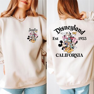 May include: Beige sweatshirt featuring a Disneyland graphic on the back with the text "Disneyland Est 1955 California". The front of the sweatshirt has a smaller graphic with the name "Jessica" and Disney characters.