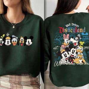May include: Dark green sweatshirt featuring a line of Disney characters including Goofy, Mickey Mouse, Donald Duck, and Pluto. The back of the sweatshirt displays a Disneyland graphic with the text "Where dreams come true."