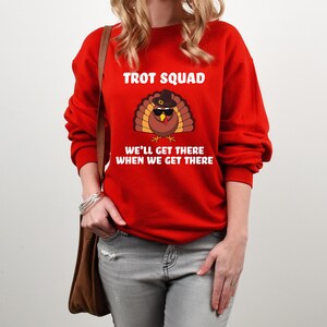 May include: A red sweatshirt featuring the words "TROT SQUAD" and a cartoon turkey wearing sunglasses and a pilgrim hat. The text "WE'LL GET THERE WHEN WE GET THERE" is below the turkey.