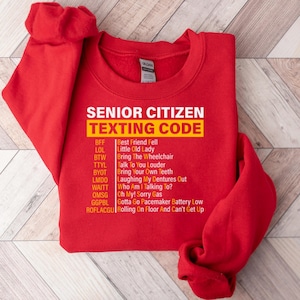 May include: Red sweatshirt with a yellow graphic that says "Senior Citizen Texting Code" and lists common texting abbreviations and their meanings.