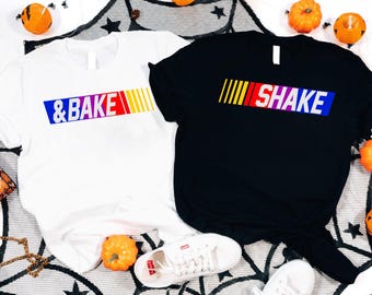May include: Two t-shirts, one white and one black, with colorful text graphics. The white shirt says "& BAKE" and the black shirt says "SHAKE". The shirts are on a Halloween-themed background with pumpkins and spiderwebs.