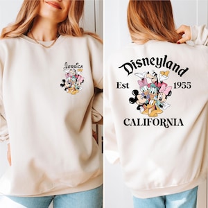 May include: Beige sweatshirt featuring a Disneyland graphic on the back with the text "Disneyland Est 1955 California". The front of the sweatshirt has a smaller graphic with the name "Jessica" and Disney characters.