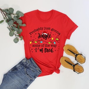 May include: A red t-shirt with the text "Probably just gonna Jingle some of the way, I'm tired." The shirt is paired with blue jeans and tan sandals with black straps. A sprig of green leaves is also present.